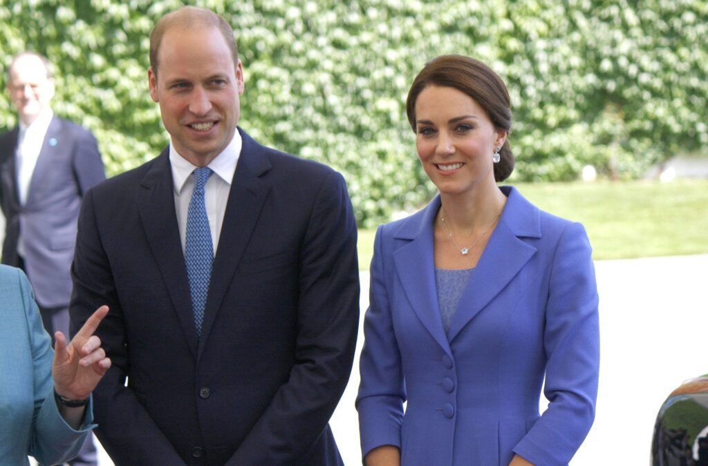 青いネクタイをした暗いスーツを着たウィリアム王子は、青いスーツを着たケイト・ミドルトンと一緒に立っています